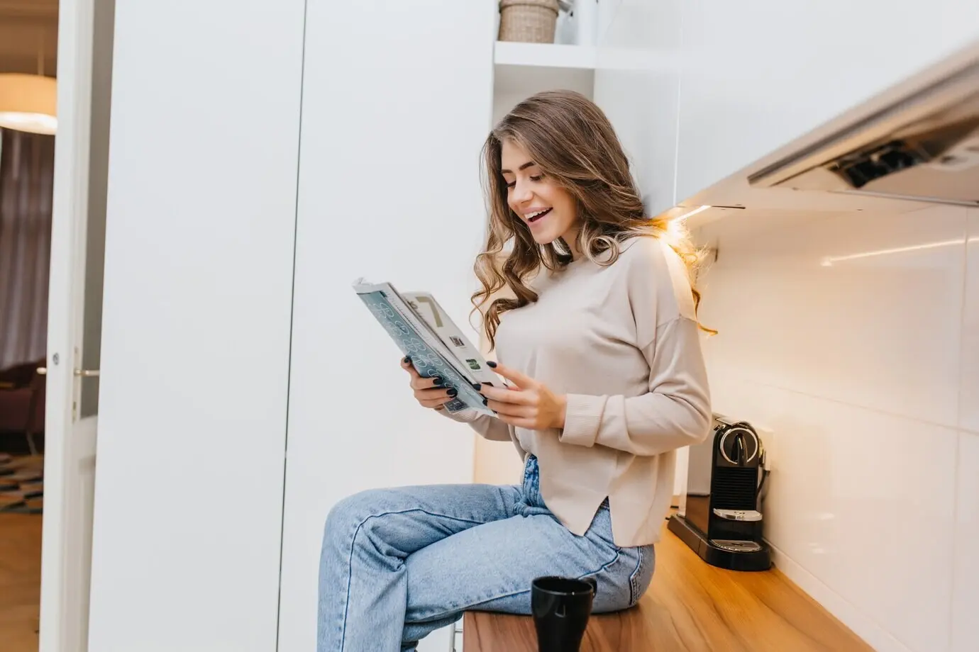 Elegantes Mädchen mit langen Haaren, das mit überschlagenen Beinen in der Küche sitzt und lächelnd Nachrichten liest.